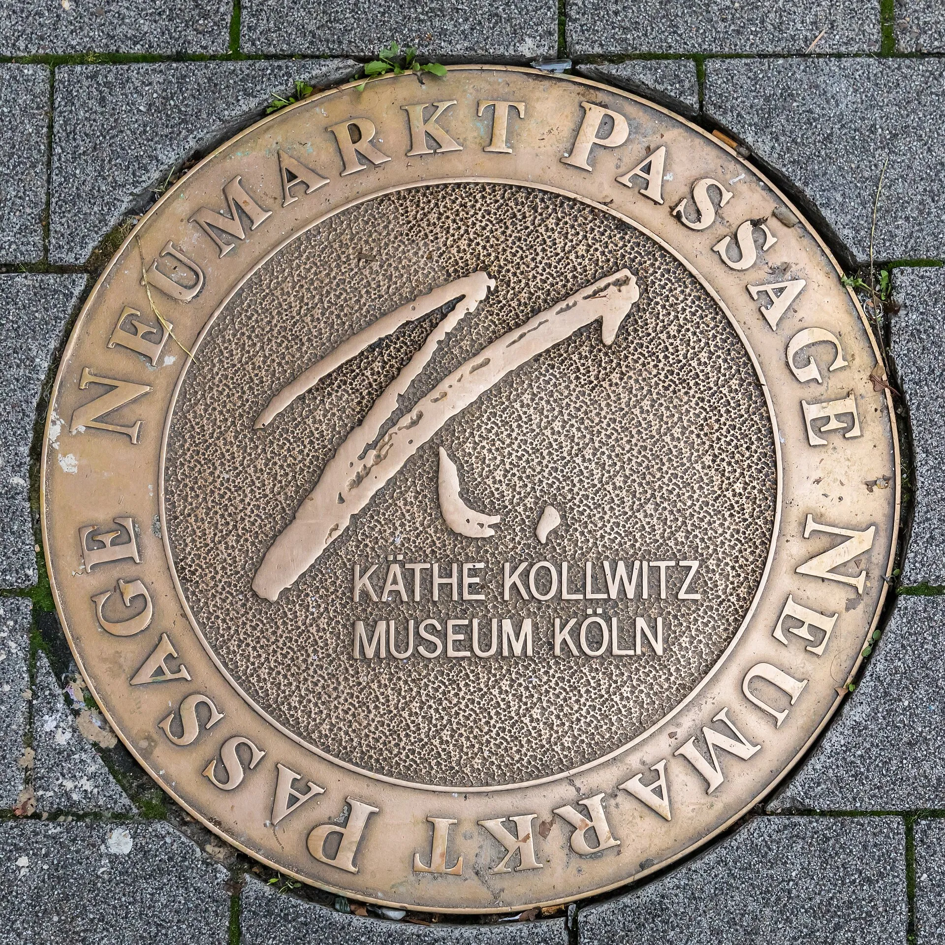 Plakette in Form und Größe einer Schachtabdeckung mit Werbung für das Käthe-Kollwitz-Museum Köln: Ort: Gehweg Neumarkt vor dem Museum