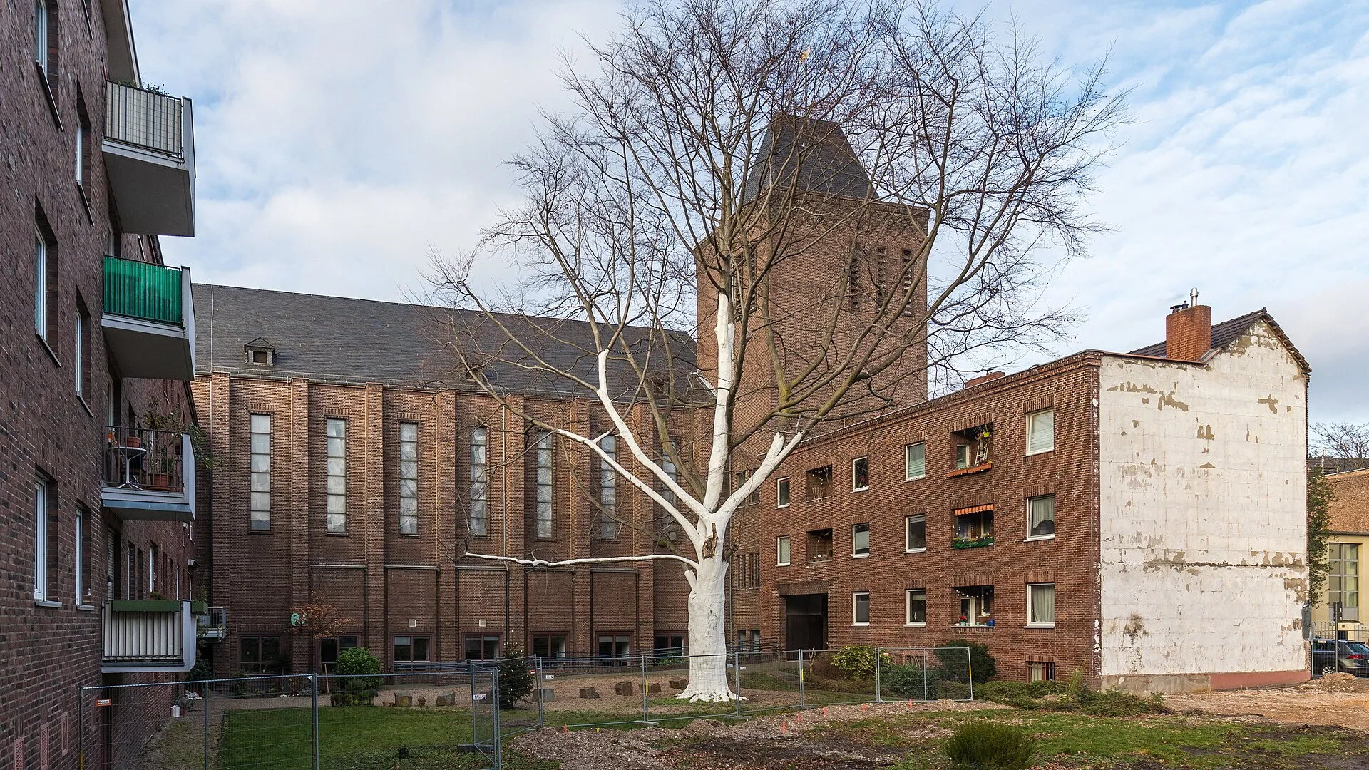 St. Karl Borromäus, Köln-Sülz. Ansicht von der Palanterstraße. Rechts das Wohnhaus Redwitzstraße 87. In der Mitte ein weiß angestrichener Baum.